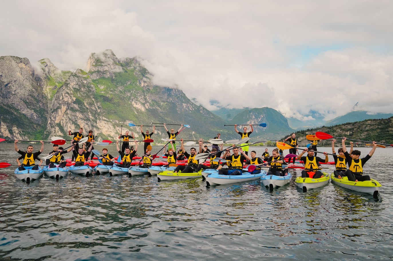 Gruppo in kayak sul Lago di Garda con le pagaie alzate davanti alle montagne